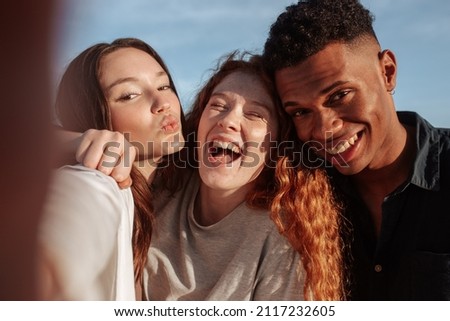 Cheerful friends taking a group selfie outdoors. Three multicultural friends posing for the camera while hanging out together on a sunny day. Group of young people making happy memories together.