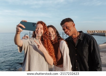 Sea selfies with friends. Group of cheerful multicultural friends taking a group photo while hanging out next to the sea. Three carefree young people making happy memories together.