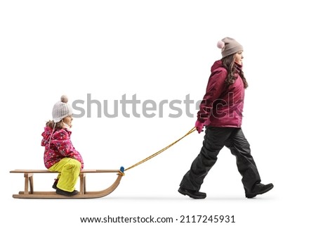 Older girl in winter clothes pulling a little girl on a sled isolated on white background