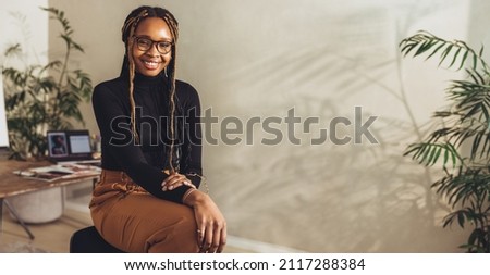 Young female freelancer smiling at the camera while working on a new project in her home office. Happy young photographer sitting on a chair with her creative desk in the background. Royalty-Free Stock Photo #2117288384