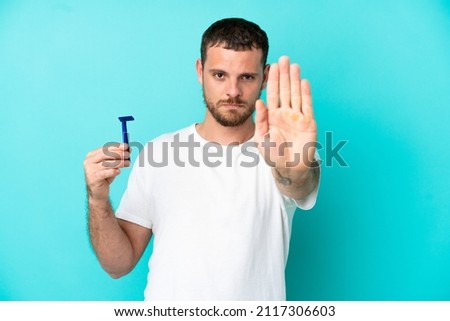 Brazilian man shaving his beard isolated on blue background making stop gesture