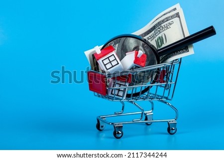 Toy model house, magnifying glass,money and keys in a shopping cart. Rent apartments, Real Estate and buying a house idea. Blue background