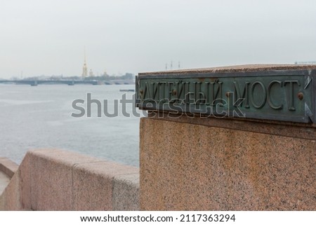 Close-up view of plaque with Russian name of Liteyny Most (Liteyny Bridge) at cold cloudy day, defosused Neva panorama at background. Bronze and granite. Copy space for text. St.Peterburg, Russia Royalty-Free Stock Photo #2117363294