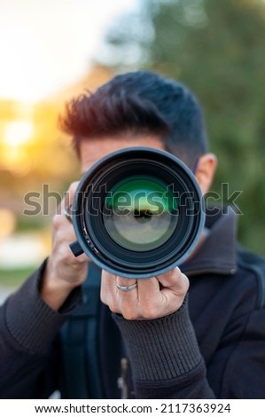 Young latin person pointing directly at you with his camera lens. Photographer taking pictures. Vertical shoot on natura. Person pointing a camera lens at you
