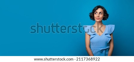 Wide banner photo of a gorgeous young woman in a blue dress, who is looking in the camera and smiling, while standing and posing isolated on blue background