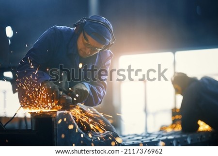 Caucasian man in protective clothes, glasses and gloves welding metal construction on factory. Professional locksmith making some details from steel at workshop.  Royalty-Free Stock Photo #2117376962