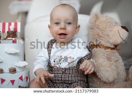 Portrait of infant little smiling baby wearing square overall, celebrating happy birthday party. Presents and gifts with cake, decorated by bears cartoons. Big teddy bear near chair.Indoor apartment.