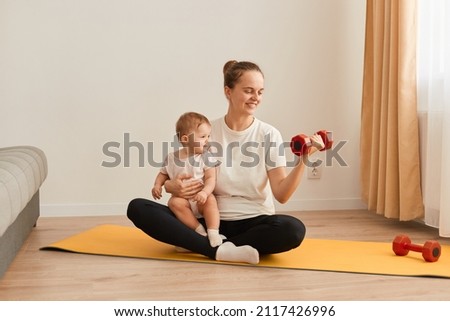 Horizontal shot of mother with baby doing gymnastics and fitness exercises at home, people sitting on yoga mat near sofa, woman holding infant daughter and training biceps and triceps with dumbbell.