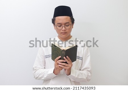 Portrait of young Asian muslim man reading and reciting Holy book of Quran seriously. Isolated image on white background Royalty-Free Stock Photo #2117435093