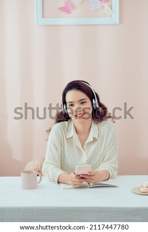 Image of serious beautiful woman using wireless headphones and cellphone while sitting at table