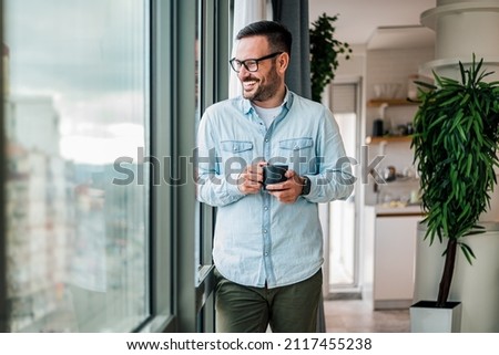 Smiling young adult businessman holding drinking coffee cup in office entrepreneur looking through window while standing in modern workplace man taking a time off or taking a brake from hard work Royalty-Free Stock Photo #2117455238