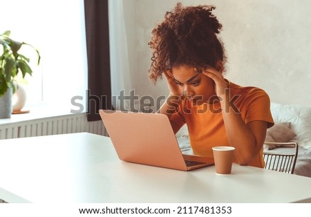 Tired flustrated African descent young girl sitting at desk in front of laptop while irritably looking at computer screen with hands holding her head at temples. Emotional side of freelancing Royalty-Free Stock Photo #2117481353