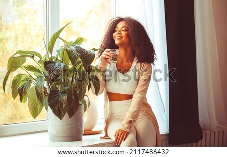 Young positive african american woman sitting on windowsill with glass of pure mineral water and looking through window with smile, drinking aqua in morning, starting new day with healthy habits Royalty-Free Stock Photo #2117486432