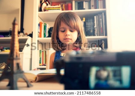 Trendy little girl in summer casual wear and white hat is recording herself with a camera at home. On the desk there is a metal souvenir of Eiffel tower. White library on background. Little blogger.