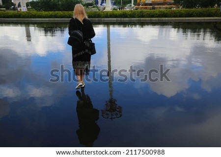 A woman walks on wet asphalt after a rain. The sky is reflected in puddles on the asphalt. It seems that the woman is walking across the blue sky.