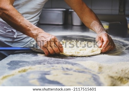 The Italian pizza maker prepares a margherita pizza by rolling out the dough with mozzarella on the work surface before cooking in the stone oven Royalty-Free Stock Photo #2117515601