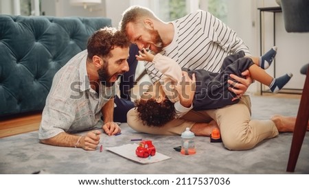Loving LGBTQ Family Playing with Toys with Adorable Baby Boy at Home on Living Room Floor. Cheerful Gay Couple Nurturing a Child. Concept of Diverse Childhood, New Life, Parenthood. Royalty-Free Stock Photo #2117537036