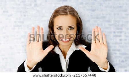 Smiling business woman in black suit showing stop gesture, on white bricks loft wall background. Businesswoman holding raising open hands, palms.