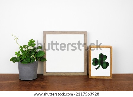 Mock up wood frame with St Patricks Day decor on a wood shelf. Shamrock plant and plaid fabric wood sign. Square frame against a white wall. Copy space.