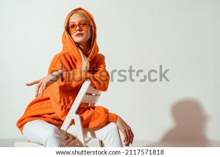 Fashionable confident blonde woman wearing trendy orange sweatshirt, color sunglasses, posing on white background. Copy, empty space for text Royalty-Free Stock Photo #2117571818