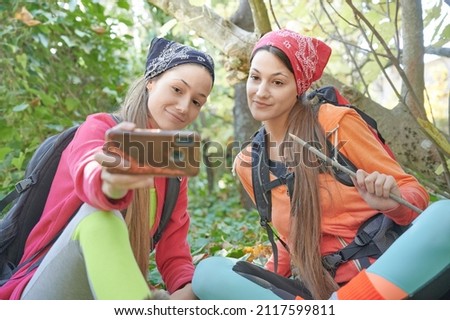 A couple of young college women enjoy a break in the shade of the trees on a sunny day of hiking and take the moment to take a selfie.