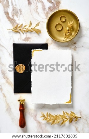 Luxury wedding stationery set. Blank invitation card mockup with black envelope, wax seal stamp, golden rings, floral leaves on marble gold desk table. Flat lay, top view.