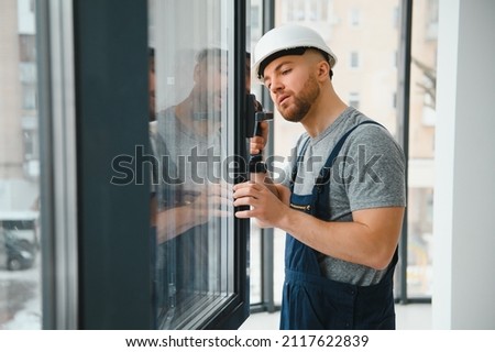 Workman in overalls installing or adjusting plastic windows in the living room at home Royalty-Free Stock Photo #2117622839