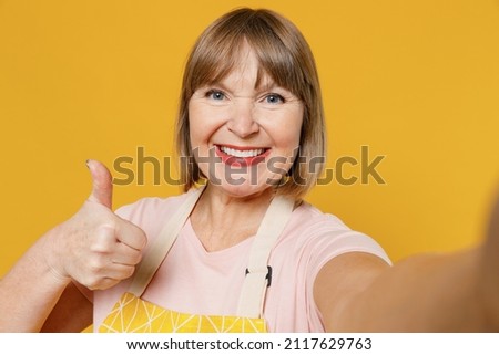 Close up smiling fun elderly housekeeper housewife woman 50s in orange apron doing selfie shot pov on mobile phone show thumb up isolated plain on yellow background. People household lifestyle concept