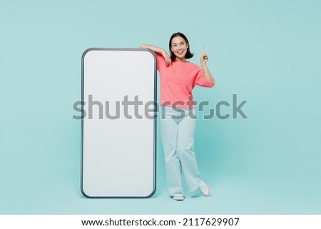 Full body young woman of Asian ethnicity in pink sweater stand near big mobile cell phone with blank screen workspace area point finger up with new idea isolated on pastel plain light blue background Royalty-Free Stock Photo #2117629907