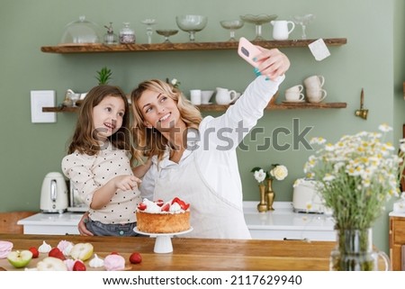 Happy chef cook baker mom woman in white shirt work baby girl helper do selfie shot mobile cell phone show pie at kitchen table home Cooking food process concept Mommy little kid daughter prepare cake