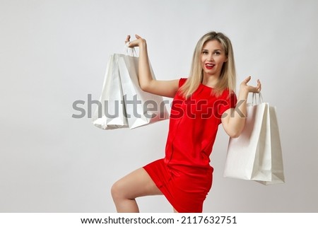Picture of a happy young blonde in a red summer dress posing with shopping bags and looking at the camera on a white background. Beautiful customer bit her tongue from the discounts she saw