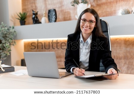 Portrait of a successful, young adult business woman, broker, financial consultant, sales manager, sit in a modern office, in stylish business clothes, working with papers, looks at camera, smiling Royalty-Free Stock Photo #2117637803