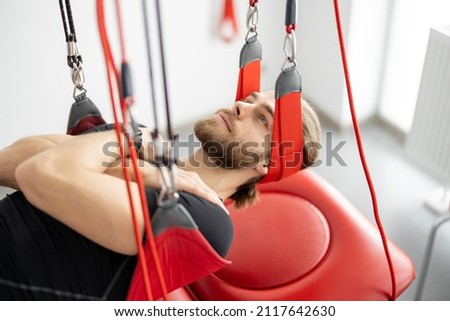 Male patient hanging on suspensions at rehabilitation center. Therapeutic exercises and neuromuscular activation on red cord slings Royalty-Free Stock Photo #2117642630