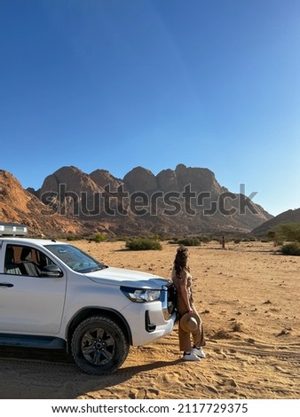 Young woman in overalls with hat stands near car in desert. SUV offroad auto vehicle white with camping equipment and rooftop tent. Safari travel. Spitzkoppe mountain in Africa, Namibia.