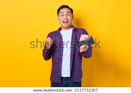 Excited young Asian man in plaid shirt holding wallet full of money banknotes isolated on yellow background Royalty-Free Stock Photo #2117752307