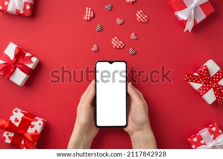 First person top view photo of st valentine's day decor young woman's hands holding smartphone checkered hearts red and white gift boxes on isolated red background with empty space