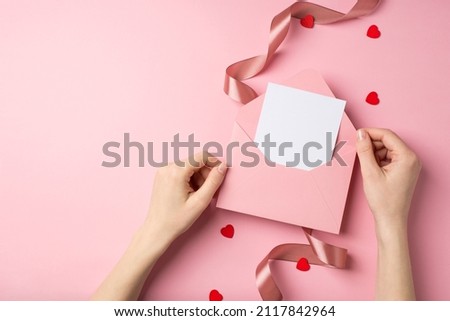 First person top view photo of valentine's day decor female hands holding pink envelope with letter red heart shaped confetti and satin curly ribbon on isolated pastel pink background with copyspace