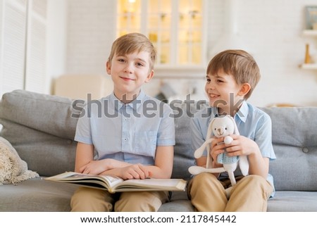 Two boys sitting on couch are reading book. Older brother reads interesting stories to his younger brother, concept friendship, intelligence, interaction, younger brother holds toy rabbit in his hand
