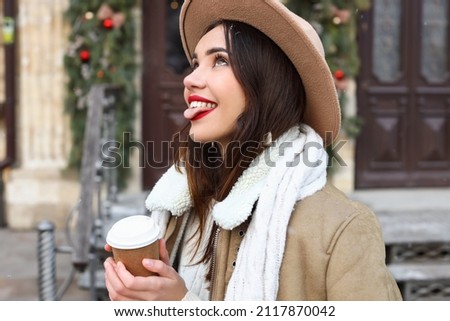 Young fashionable woman with paper cup catching snowflakes with tongue on winter day