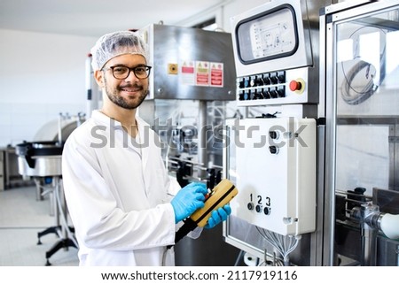 Portrait of technologist or worker in sterile white clothing standing by automated industrial machine in pharmaceutical company or factory.