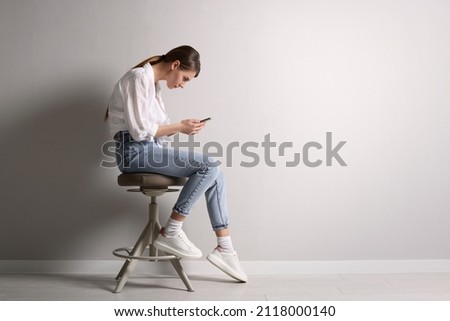 Woman with bad posture using smartphone while sitting on stool near light grey wall indoors, space for text Royalty-Free Stock Photo #2118000140