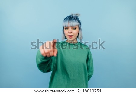 Positive teen girl with blue hair points a finger at the camera and smiles on a blue background