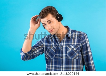 portrait of stylish attractive handsome young man listening to music on wireless headphones having fun modern style happy emotional mood isolated on blue background wearing checkered shirt