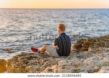 A young blond man stands on the seashore in front of a bright orange sunset. Romantic seascape