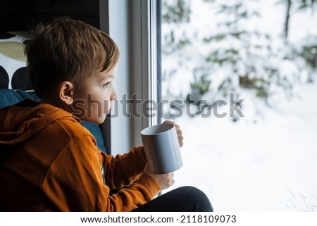 A cute boy drinks warm cocoa and sits by the window. Winter landscape outside the window. Schoolchildren on vacation. comfort and tranquility. High quality photo