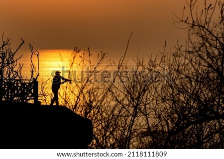 Silhouette shot of man in hat fishing with fishing rod on cliffs at sunset