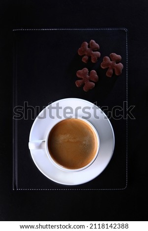 A white coffee cup and a black leather-bound notebook.Flat lay.Top view.Copy space.Food and drinks.Coffee break.Cookies in the shape of a clover.Cookies are out of focus.