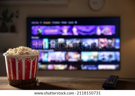 Close up of bowl of popcorn and remote control with tv works on  background. Evening cozy watching a movie or TV series at home.  Royalty-Free Stock Photo #2118150731