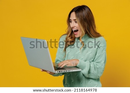 Young surprised overjoyed excited amazed woman 30s wearing green knitted sweater hold use work on laptop pc computer isolated on plain yellow color background studio portrait. People lifestyle concept