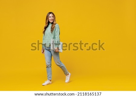 Full body young fun woman 30s in green knitted sweater hold closed pc use work on laptop computer look aside on copy space area isolated on plain yellow background studio. People lifestyle concept.
